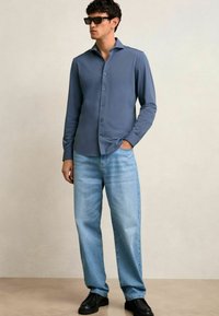 Hemd in Schieferblau, langärmelig, glatte Textur. Kombiniert mit hellblauen geraden Jeans und schwarzen Schuhen. Dunkle Sonnenbrille getragen.