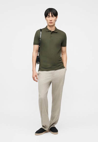 Άνδρας που φοράει πουκάμισο πόλο ελιάς, μπεζ παντελόνι λινό, μαύρα εσπαντρίγιες και κρατάει μια μικρή μαύρη τσάντα ώμου σε γκρι φόντο.