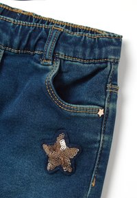 Gros plan sur une poche de jean en denim bleu avec ceinture élastique, rivet en étoile doré et un patch étoile en sequins brillants en dessous de la poche.