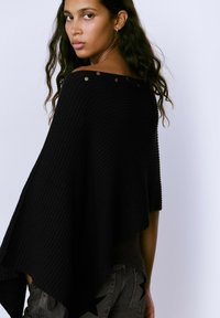 Poncho negro de canalé con escote amplio y botones decorativos en el hombro. Textura suave, forma fluida y dobladillo asimétrico.