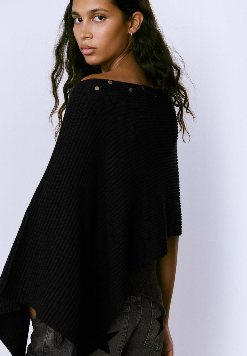 Poncho negro de canalé con escote amplio y botones decorativos en el hombro. Textura suave, forma fluida y dobladillo asimétrico.