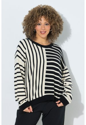 Kvinde med krøllet hår iført en sort og hvid stribet sweater med lodrette og vandrette mønstre, stående mod en hvid baggrund.