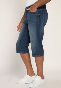 Pantalons capri en denim bleu foncé avec une coupe droite, des coutures visibles et cinq poches. Assortis à des chaussures de sport blanches sur un fond clair.