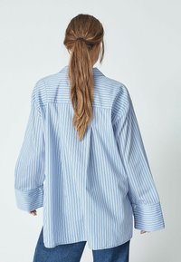 Blue striped shirt with a loose fit, featuring wide sleeves and a button-down back. Made of smooth fabric, it has a relaxed design.