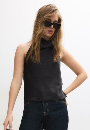 Donna con capelli biondi e lunghi, che indossa una canottiera nera, jeans scuri e occhiali da sole rettangolari neri, in piedi davanti a uno sfondo bianco.