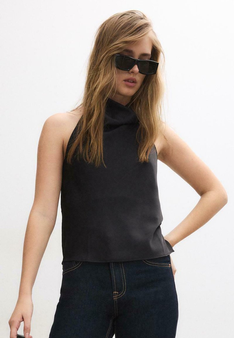 Žena s dlhými blond vlasmi, oblečená v čiernom tielku bez rukávov, tmavých džínsoch a obdĺžnikových čiernych slnečných okuliaroch, stojaca na bielom pozadí.