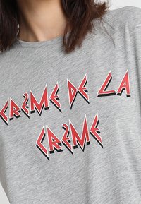 Grå T-shirt med fed rød og sort stiliseret tekst "CRÈME DE LA CRÈME" båret af en person med skulderlangt brunt hår.
