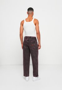 Maroon denim jeans with a loose fit, featuring a buttoned waistband, back pockets, and subtle faded texture. Worn with a white tank top.