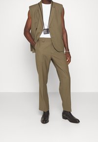 Olijfgroen op maat gemaakt pak met mouwloze jas, slim-fit broek en wit T-shirt met ronde hals. Bevat een minimalistische zwarte crossbody tas en zwarte laarzen.