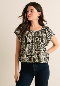 Blouse à manches courtes noire et beige à motifs avec un col froncé et une coupe loose, associée à un jean sombre. Présente des motifs floraux et géométriques.