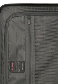 Interior de maleta de tela negra con bolsillo de malla y cierres de cremallera, que presenta una etiqueta con el logo y un sutil patrón de rayas verticales.