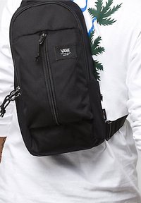 Svart Vans slingväska med en blixtlåsförsedd frontficka, justerbar rem och texturerat material. Har en logopatch på framsidan.