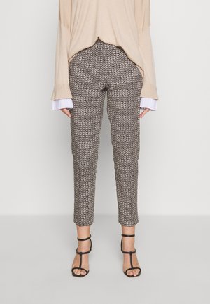 Kvinde iført tætsiddende mønstrede bukser, beige langærmet top med plisserede manchetter og sorte sandaler med hæl og remme.