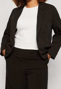 Blazer negro ajustado con manga larga, solapas muesca y cierre de un solo botón, combinado con una camisa blanca ajustada. Textura de tejido suave.