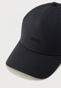 Gorra de béisbol negra hecha de tela suave, con una visera curva y detalles cosidos. "HUGO" está bordado en negro en la parte frontal.