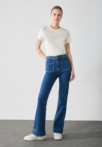 High-waisted, flared blue denim jeans with two back pockets, paired with a fitted cream T-shirt and white sneakers on a gray floor.