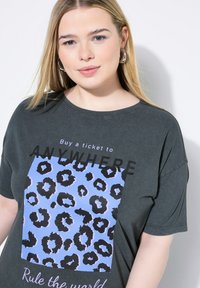 Camiseta oversized de color gris oscuro con un cuadrado azul con estampado de leopardo negro y texto blanco que dice "Compra un billete a CUALQUIER LUGAR" y "Gobierna el mundo."