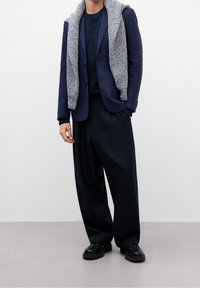 Blazer navy sobre un suéter oscuro texturizado, con una bufanda de patrón ligero apoyada en los hombros, combinada con pantalones oscuros holgados y zapatos negros.