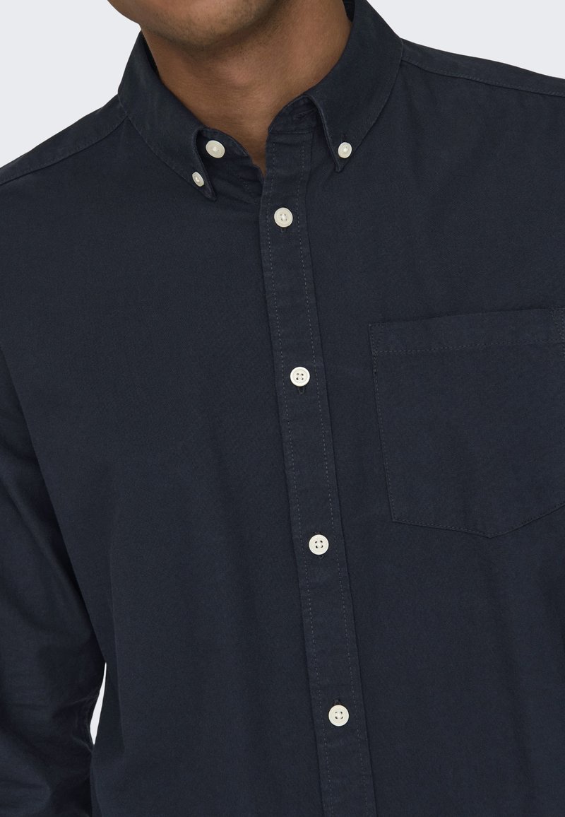 Camicia blu navy con bottoni, realizzata in tessuto dalla texture liscia; presenta un colletto appuntito, una tasca sul petto e bottoni bianchi.