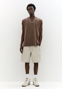 Top senza maniche marrone in un tessuto morbido, abbinato a pantaloni cargo beige. Gli accessori includono una collana in argento e sneakers bianche.