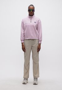 Άτομο που στέκεται φορώντας ανοιχτό μωβ φούτερ με φερμουάρ έως τη μέση, μπεζ παντελόνι cargo, γκρι αθλητικά παπούτσια και αντανακλαστικά αθλητικά γυαλιά ηλίου.