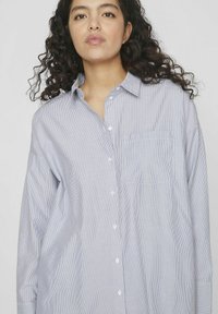 Camisa de botones a rayas en azul claro y blanco. Presenta un bolsillo en el pecho, mangas largas y un ajuste relajado con un dobladillo redondeado.