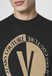 Zwarte gebreide trui met een groot beige cirkelvormig logo met de tekst "JEANS COUTURE" en een prominente "V" in het midden.