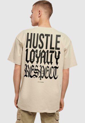 Beige színű pamut póló, nagy fekete gótikus felirattal a hátsó részén: "HUSTLE LOYALTY RESPECT", laza szabással és rövid ujjakkal.