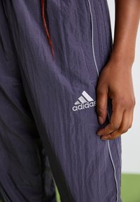 Ruka odpočíva na tmavošedých tréningových nohaviciach Adidas s bielym logom a tenkým vertikálnym pruhom.