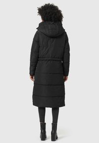 Abrigo largo negro acolchado con capucha, que presenta una cintura ajustada, textura acolchada horizontal y un diseño hasta la rodilla. Usado sobre pantalones negros y botas.