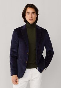 Blazer de pana de color azul marino con dos botones marrones, llevado sobre un cuello de tortuga verde oscuro, combinado con pantalones blancos. Textura suave, diseño estructurado.