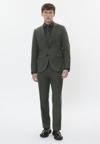 Vyras, dėvintis alyvuogių žalumos kostiumą su juostuotu juodu ryšiu, tamsia marškinėliais ir juodais elegantiškais batais, prieš baltą foną.