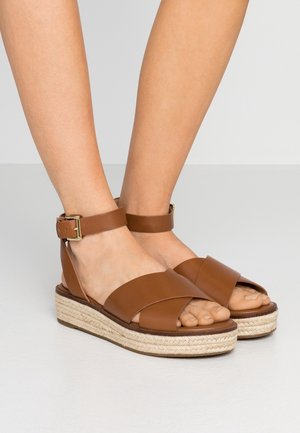 Pieds chaussés de sandales compensées en cuir marron avec des brides chevilles et des boucles dorées, posés sur une surface claire sur un fond gris clair.
