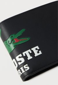 Juodas odinis piniginių su žaliu krokodilo logotipu, turinčiu raudonas burnas ir baltą užrašą. Lygus paviršius, kontrastingos spalvos ir iškilūs detalės.