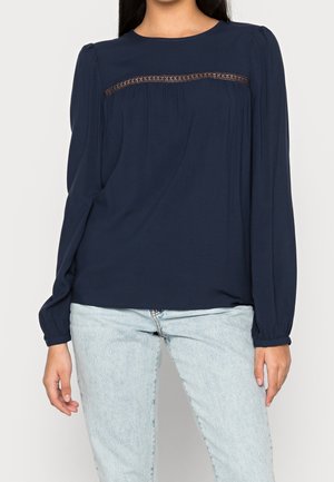 Granatowa bluzka z długim rękawem wykonana z lekkiego materiału, ozdobiona plisami i koronką na wysokości klatki piersiowej. Luźny krój, okrągły dekolt i elastyczne mankiety.