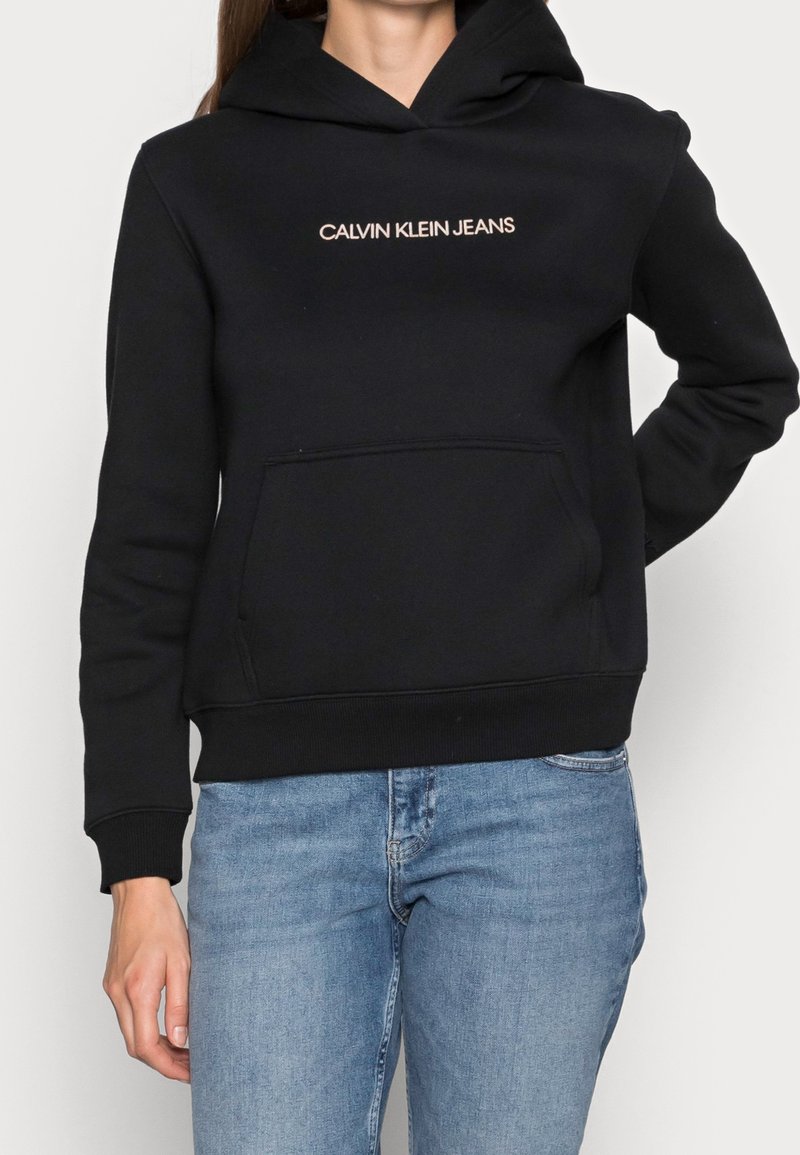 Sweat à capuche noir avec poche avant et le texte "CALVIN KLEIN JEANS" sur la poitrine, porté avec un jean bleu clair.