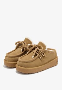 Et par tan suede slip-on sko med tykke gummisåler, runde tåspidser, snørebånd foran og hvid fleece-foring synlig ved kraverne.