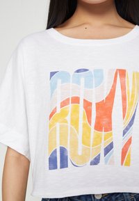 Femme portant un t-shirt court blanc avec un imprimé abstrait coloré et des manches courtes retroussées, assorti à un jean bleu.