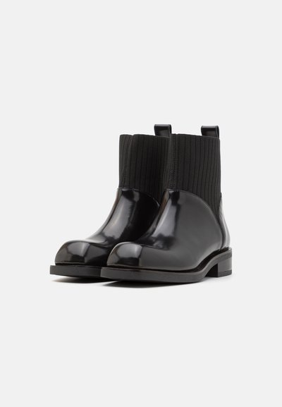 Um par de botas de tornozelo pretas com seção inferior de couro brilhante e punho superior de malha canelada, com puxadores na parte traseira, em um fundo branco liso.