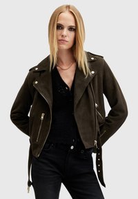 Giacca da moto in suede verde scuro con zip argento, spalline e bottoni a scatto; indossata su un top in maglia nera. Maniche lunghe.