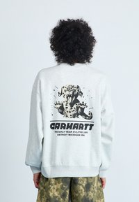 Hallikas sviiter, millel on graafiline disain, kus on mustade laikudega koer, tekst "Carhartt" ja ribatud varrukad. Kandub koos kamoflaažipükstega.