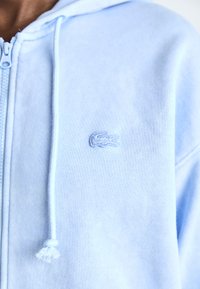 Sudadera con cremallera de color azul claro, hecha de una tela suave, que presenta un pequeño logotipo bordado en el pecho y un cordón para la capucha.