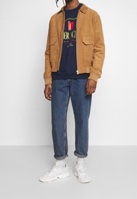 Giacca color corda con due tasche frontali sopra una felpa blu navy con una grafica. Pantaloni in denim blu a righe e scarpe da ginnastica bianche.