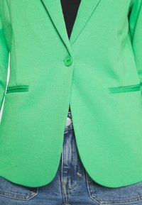 Zielona marynarka z teksturowanej tkaniny, z pojedynczym guzikiem, dwoma przednimi kieszeniami i dopasowanym krojem, noszona na jeansowych szortach.