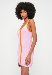 Pletena haljina u ružičastoj boji s narančastim naglascima, s rebrastom teksturom i V-izrezom. Bez rukava, s kontrastnim bočnim panelom.