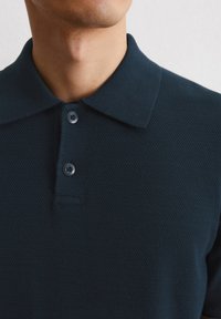 Ciemnogranatowa koszulka polo z teksturowanej tkaniny, z klasycznym kołnierzykiem i zapięciem na dwa guziki. Widoczny subtelny, tkany wzór.