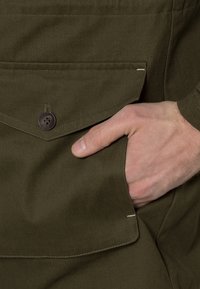 Veste vert olive en tissu de coton texturé ; comprend une poche à rabat, une fermeture par bouton marron, et une main dans la poche.
