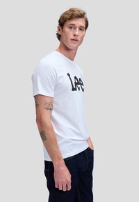 Ung mann med lys brunt hår som har på seg en hvit Lee t-skjorte og mørke jeans, stående med den ene hånden i lomma foran en ensfarget bakgrunn.