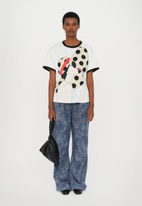 Graphic t-shirt featuring a large polka dot pattern and illustrated face. Paired with wide-leg, floral-patterned blue jeans. Black bag.