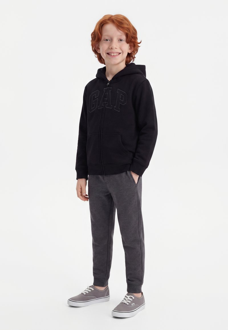 Enfant souriant aux cheveux roux portant un sweat à capuche noir zippé, un pantalon de jogging gris foncé et des baskets grises, debout avec une main dans la poche.